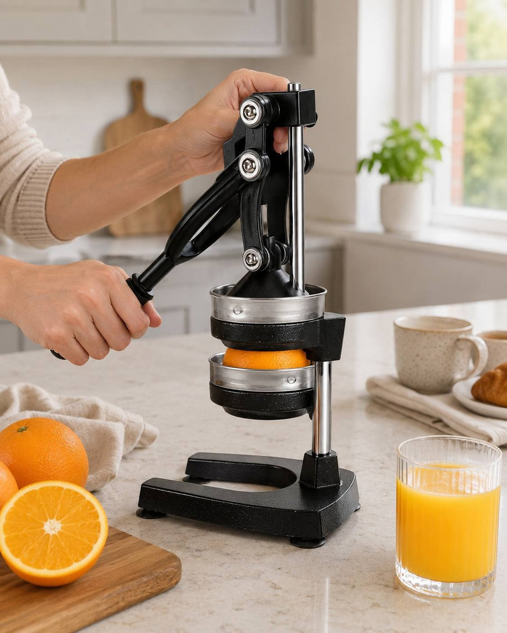A person operating a black manual lever citrus juicer on a kitchen countertop, with a halved orange placed in the metal press and a glass of freshly squeezed orange juice next to it.