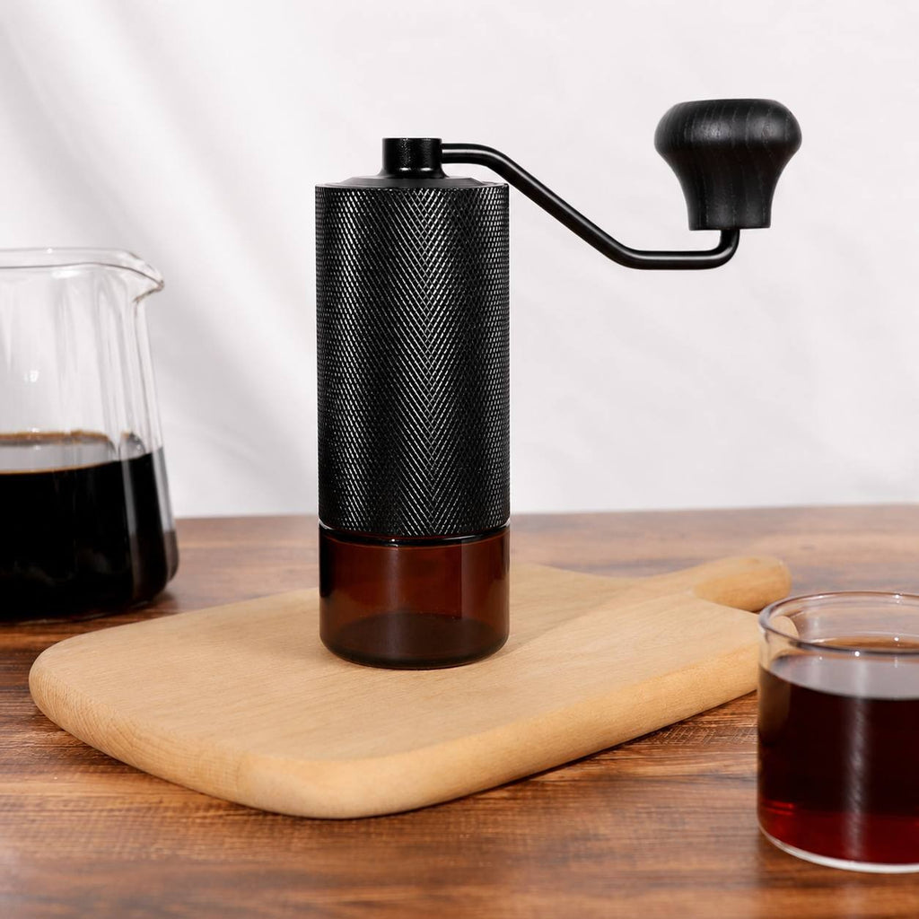 black manual coffee grinder with a knurled aluminum body and wooden crank handle standing on a wooden cutting board.