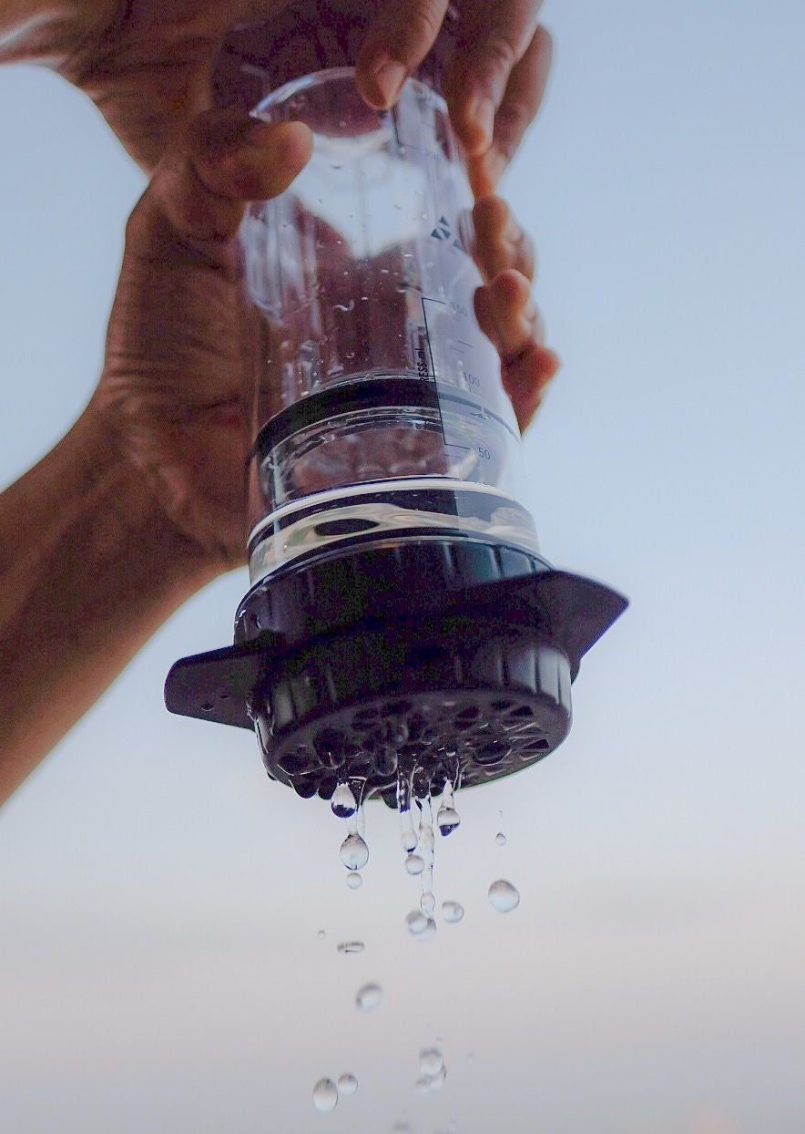 Hands holding a clear manual coffee press maker with water droplets dripping through the filter cap