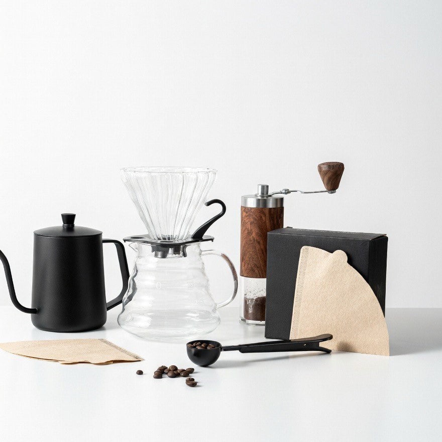 Collection of coffee-making tools including a teapot, glass carafe, grinder, and coffee beans on a white surface.