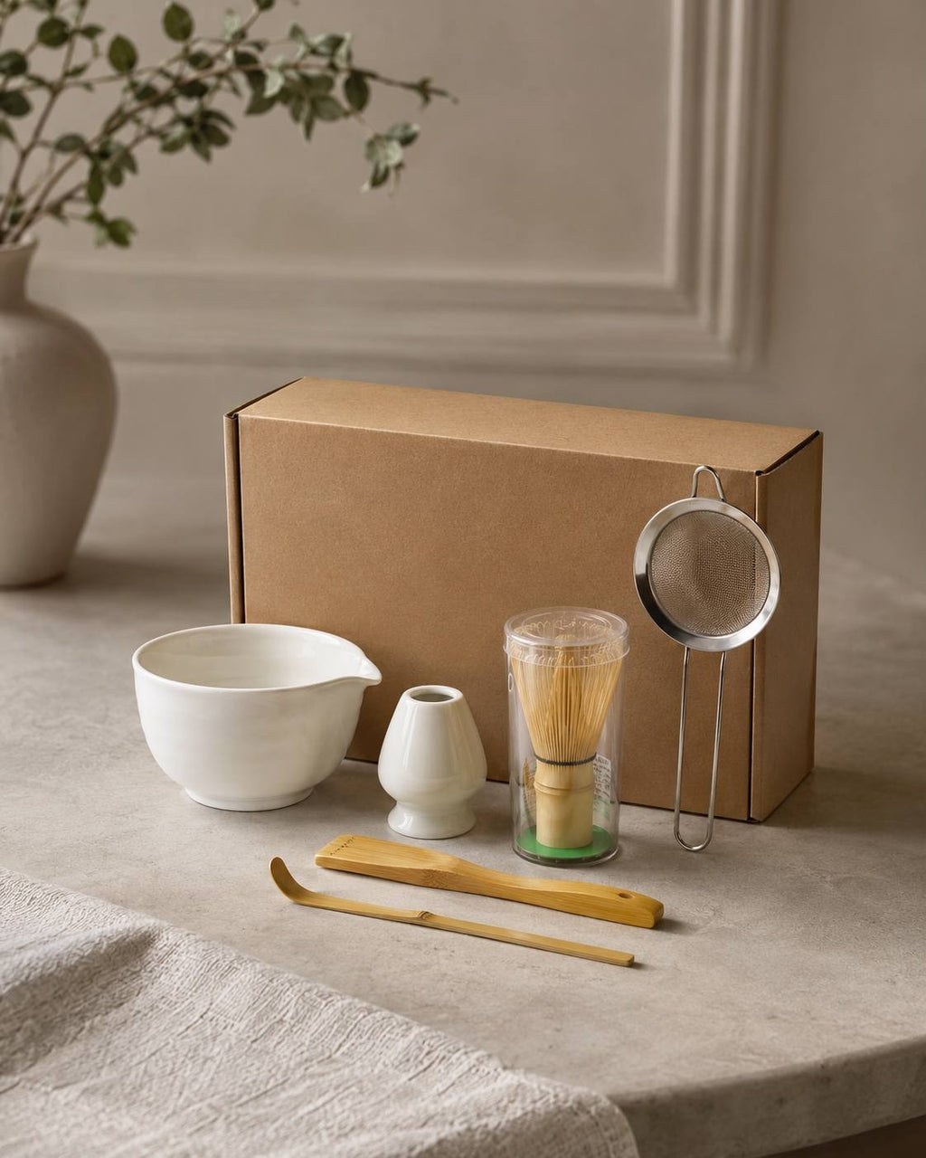 Matcha tea set featuring a white ceramic mixing bowl, whisk, scoop, and strainer on a table with a branch decoration.