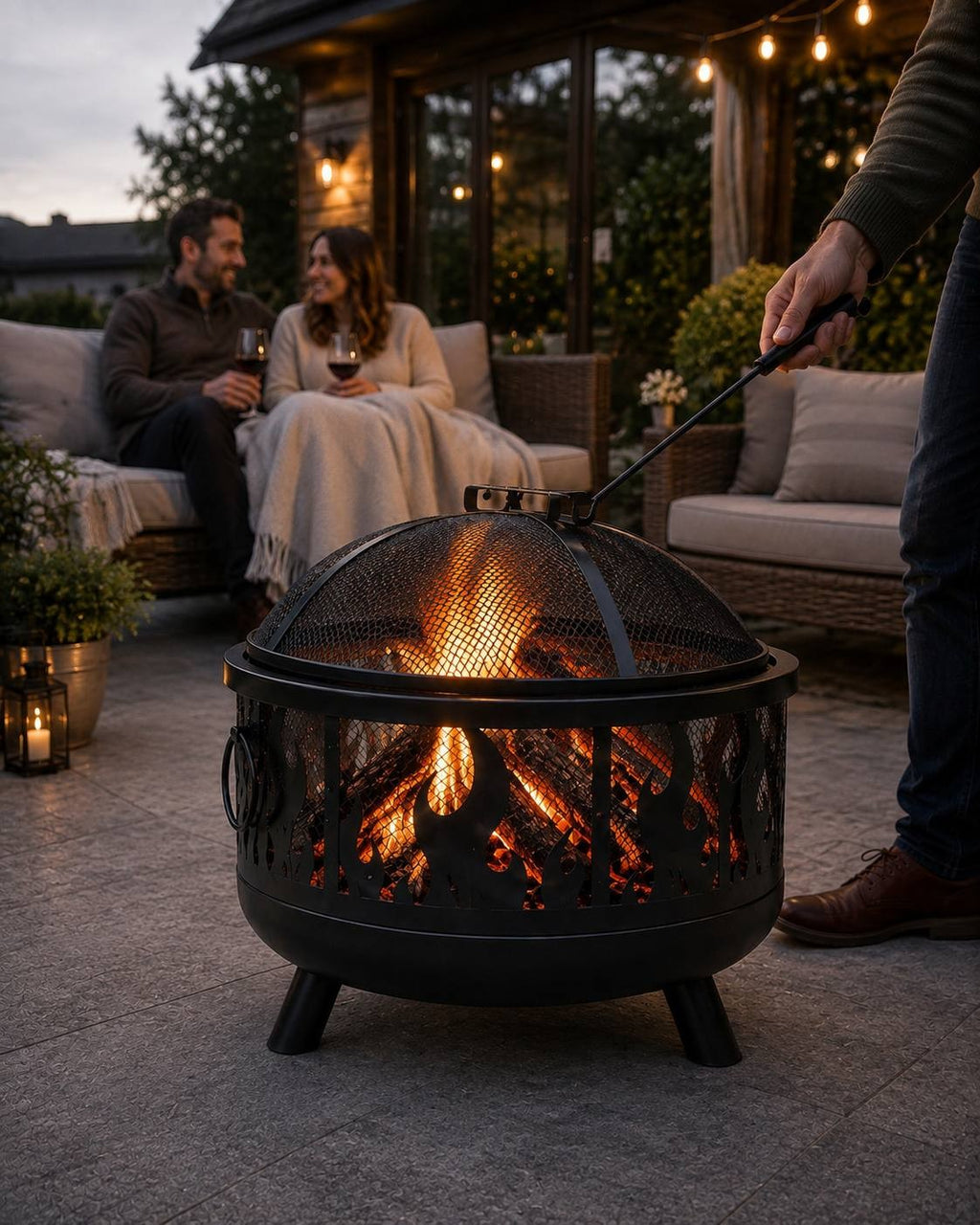 Person using a poker tool to lift the mesh spark guard lid off a black metal fire pit on an outdoor patio while a couple relaxes on a sofa in the background.
