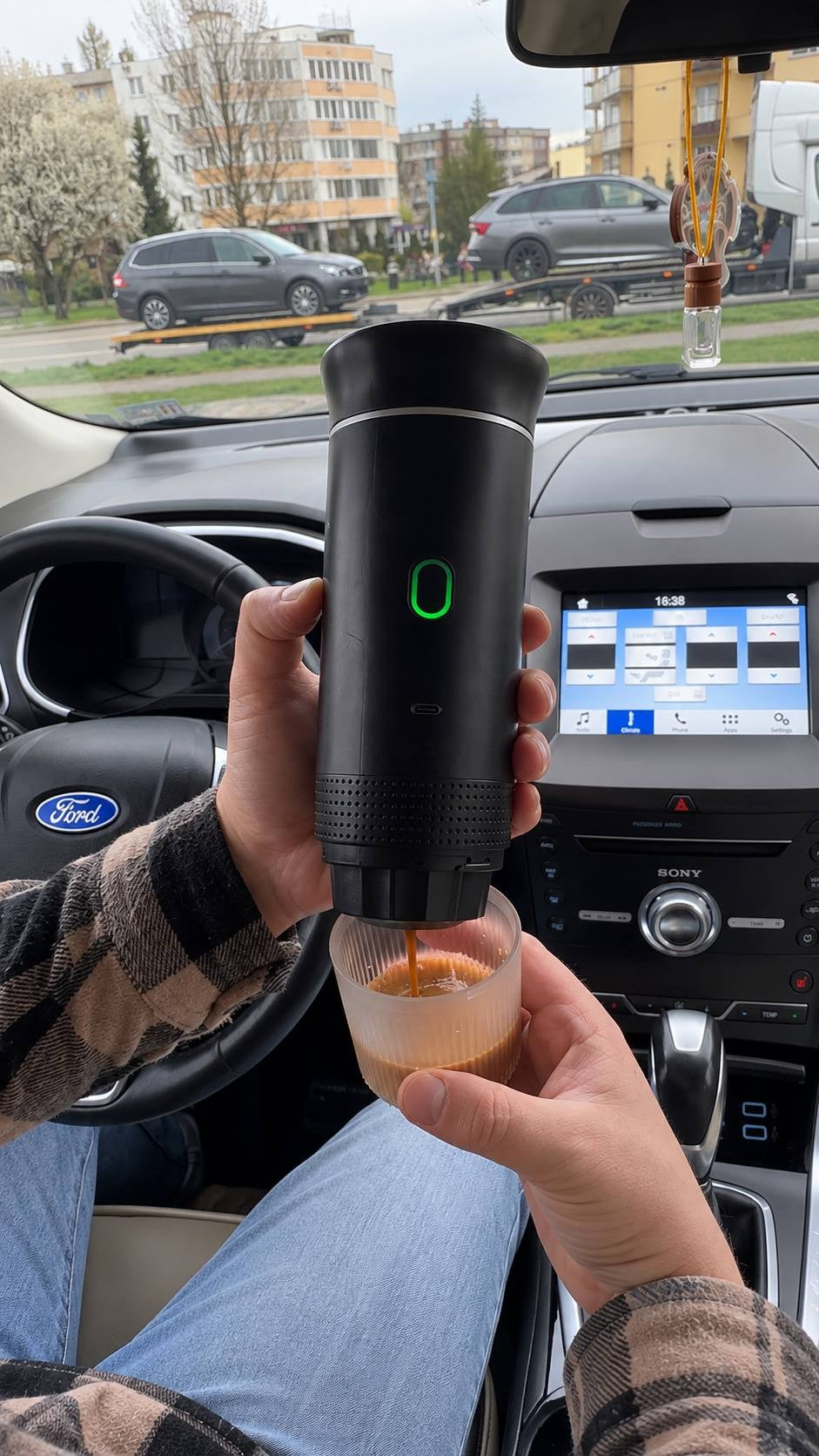 Person using a portable battery-powered espresso maker inside a Ford car, extruding rich espresso into a cup over the center console.