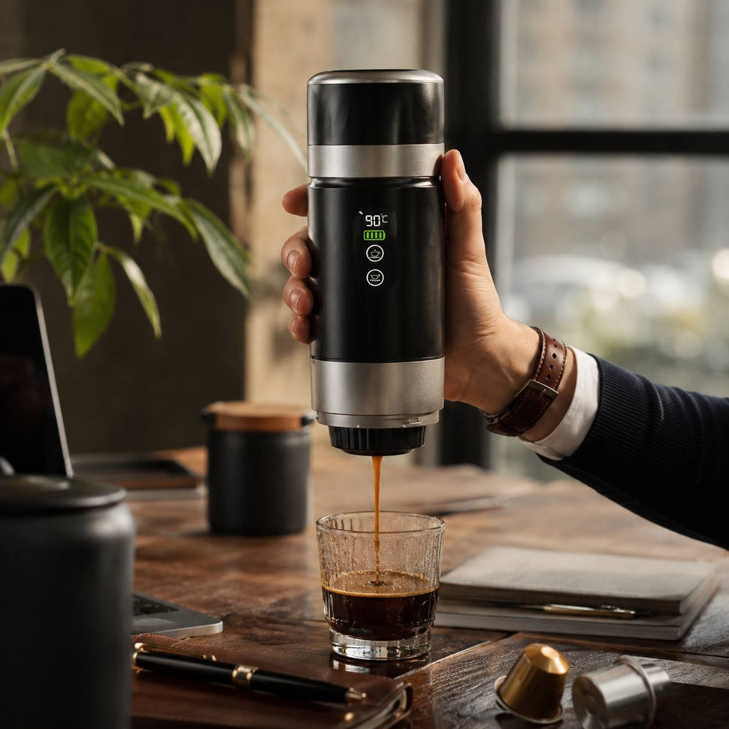 A person holding a portable electric espresso maker with a digital screen showing 90°C, brewing coffee into a glass with capsules on the wooden desk.