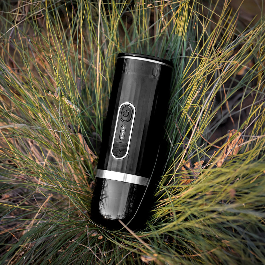 Black portable electric espresso machine lying in the green and yellow grass during a hiking trip.