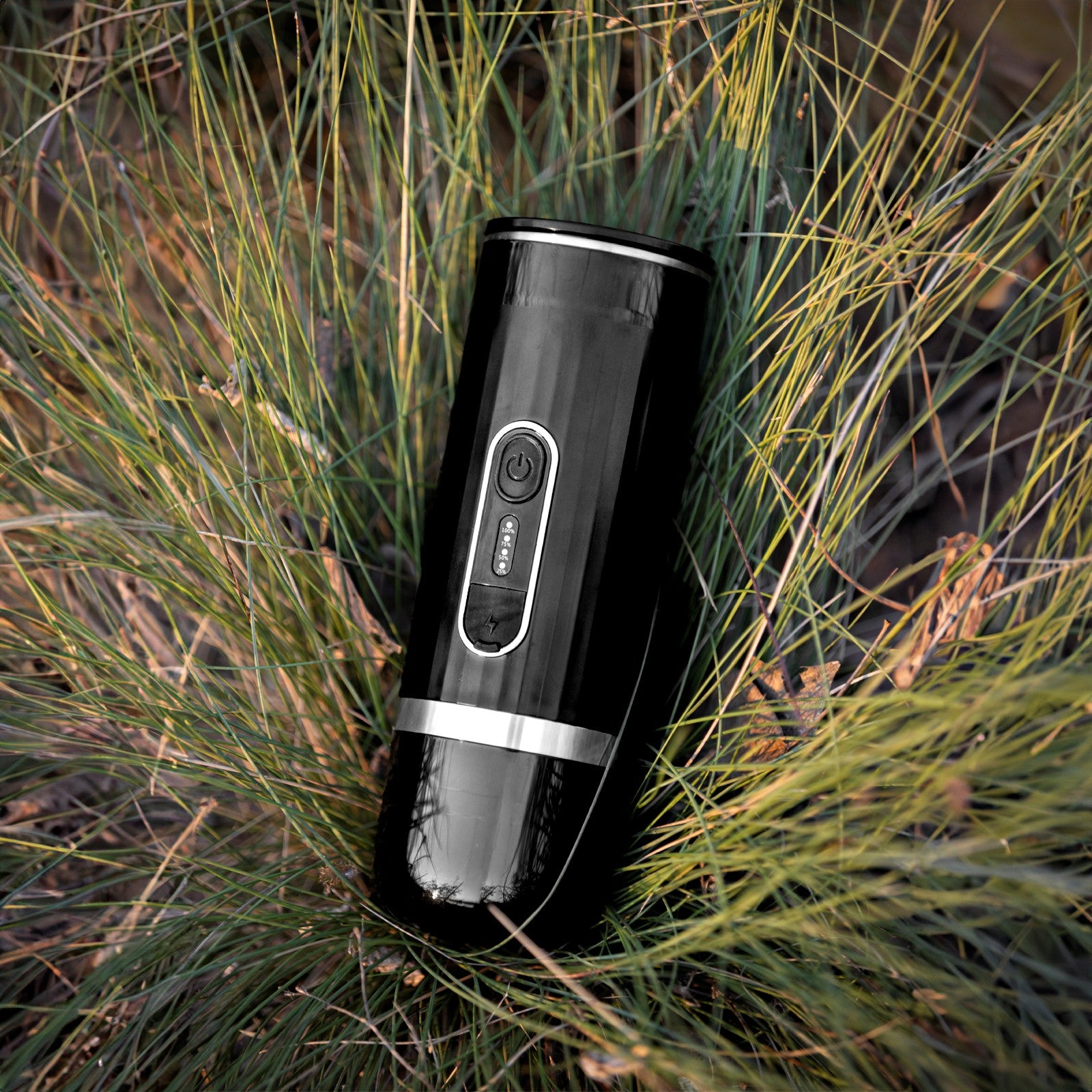 Black portable electric espresso machine lying in the green and yellow grass during a hiking trip.