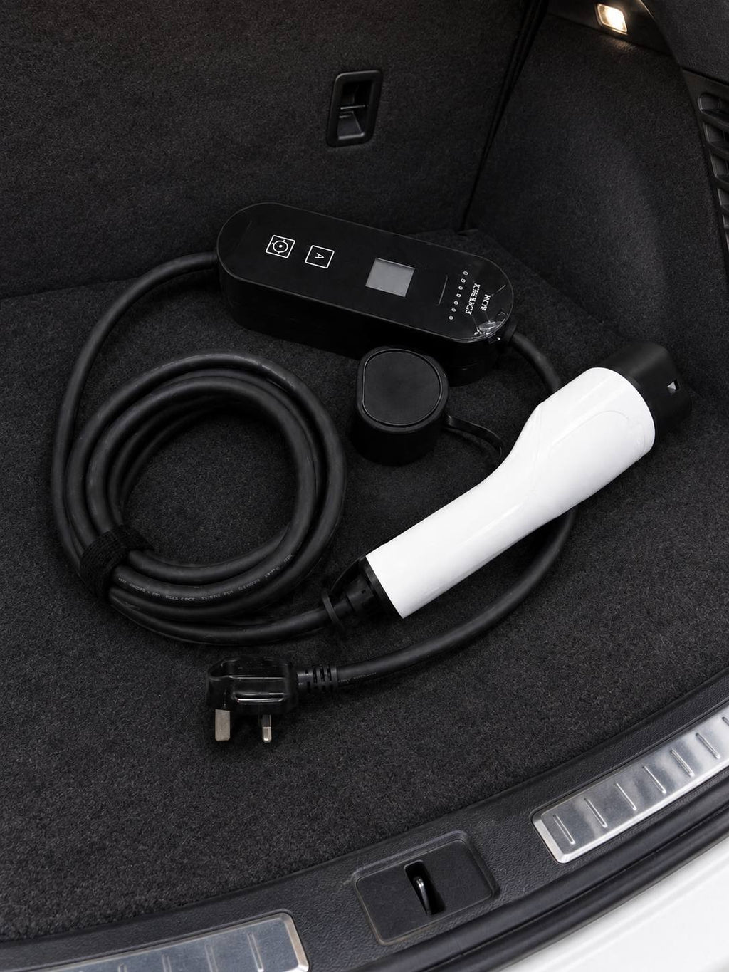 A top-down view of a portable EV charging kit neatly coiled and stored inside the carpeted trunk of an electric vehicle, including the control box, cable, and UK plug.