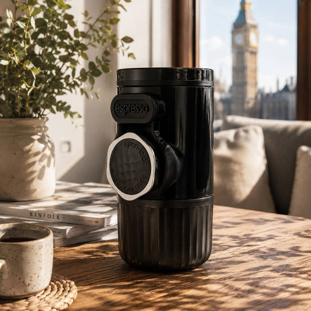 A compact black portable espresso maker standing on a wooden table next to a stack of lifestyle magazines and a fresh cup of coffee.