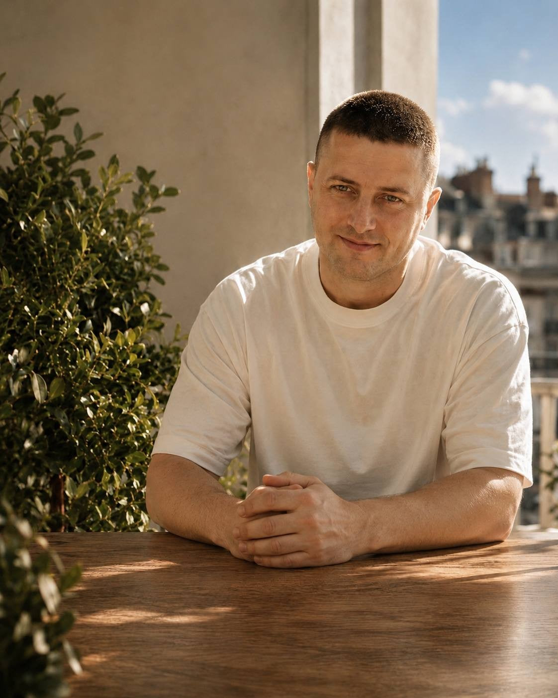 A man with short hair wearing a white t-shirt sitting at a wooden table outdoors with his hands clasped, with green plants and a cityscape blurred in the background under warm sunlight.