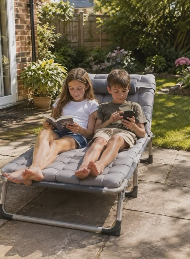A young girl reading a book and a boy using a smartphone while relaxing together on a grey padded folding sun lounger in a sunny backyard garden.