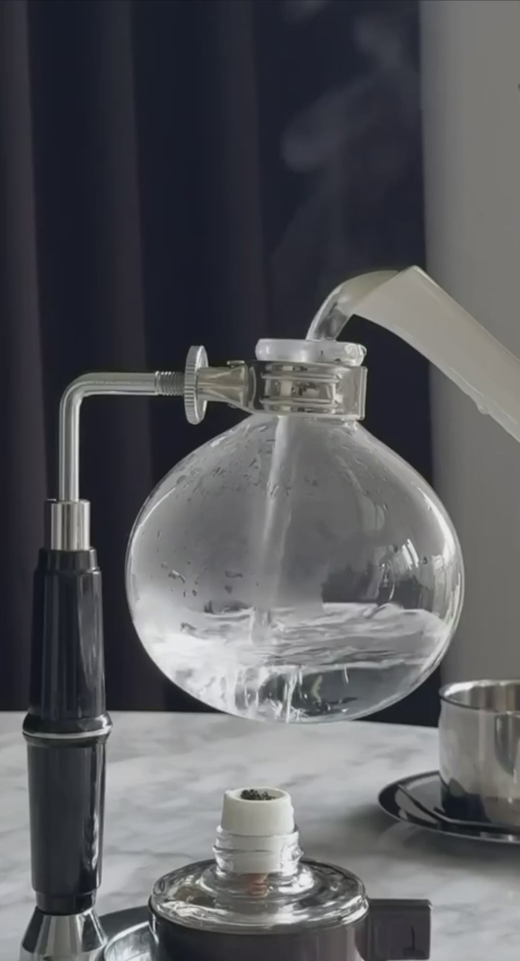 The coffee brewing process in a vacuum siphon coffee maker: a hand places the upper glass chamber onto the lower spherical vessel where water is being heated, followed by the stages of boiling and coffee extraction over an open flame.