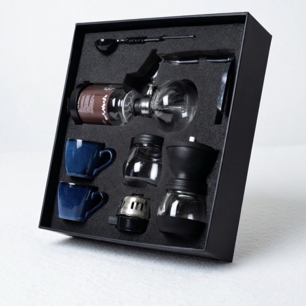 Coffee brewing set with blue cups, glass carafe, and black accessories in a black box on a white background