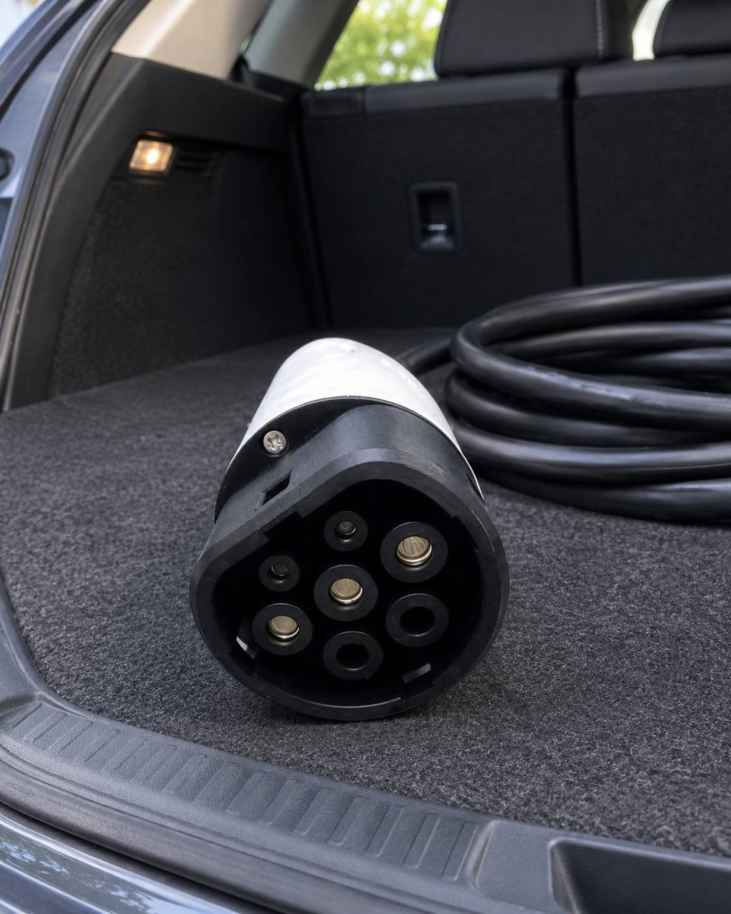 Close-up of a white and black Type 2 EV charging plug resting inside the carpeted trunk of a car next to a coiled black charging cable.