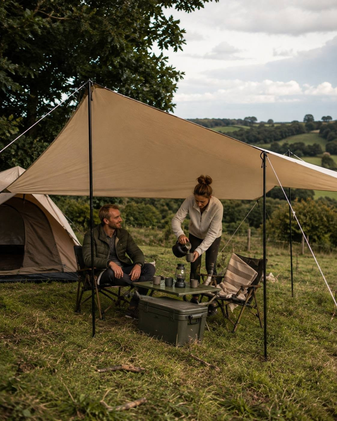 A woman pouring hot coffee from a kettle into a mug at a campsite setup under a beige canopy tarp next to a tent.