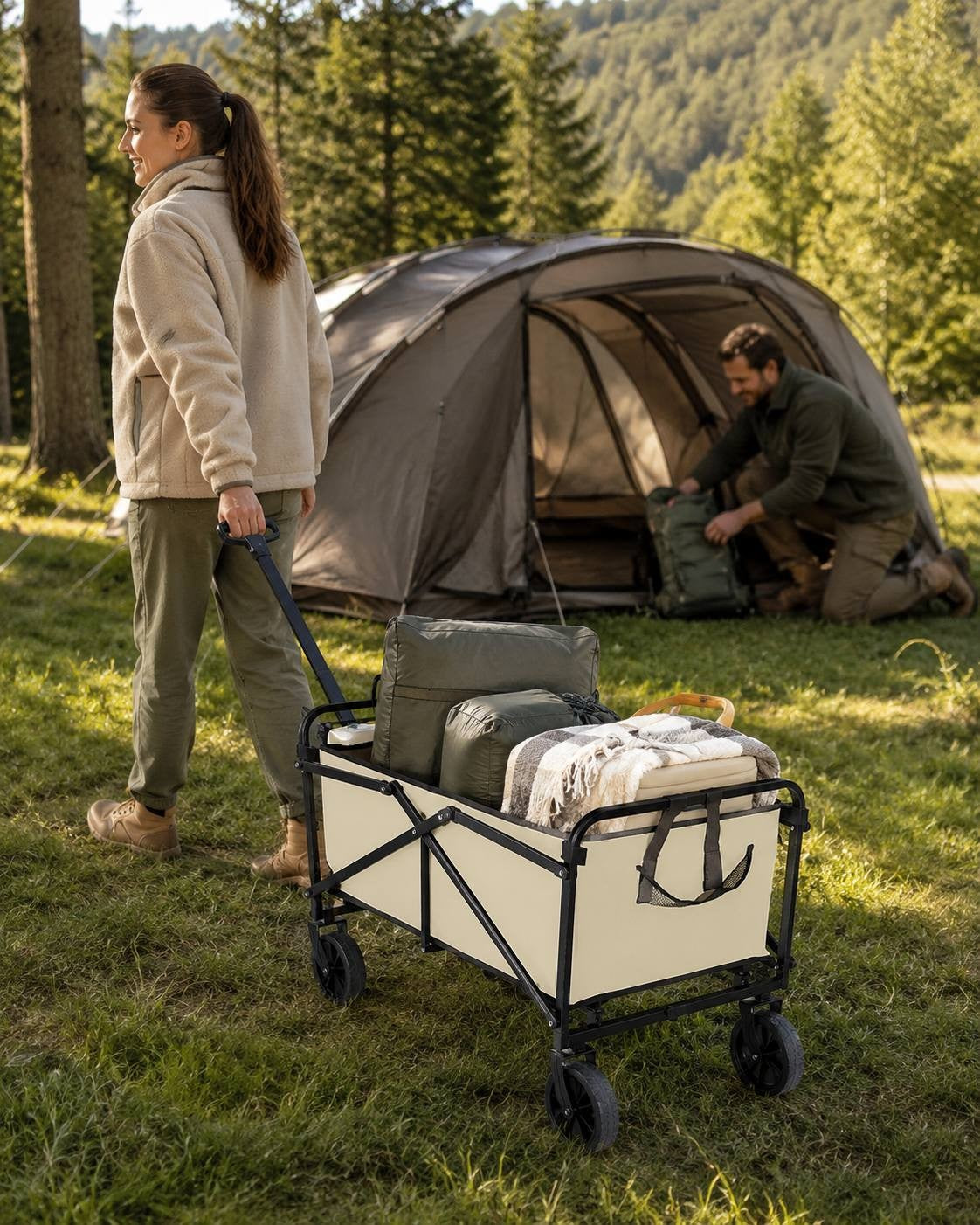 Woman in fleece jacket pulling a beige utility cart filled with sleeping bags and blankets along a grassy trail at a forest campsite with a tent.