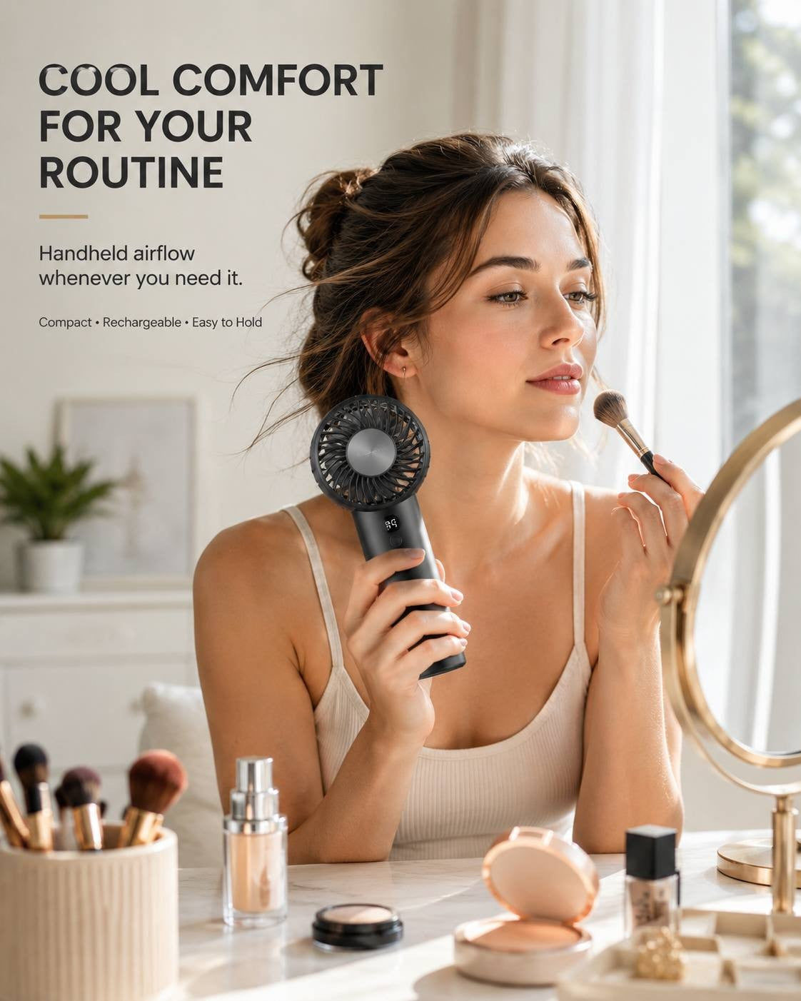A woman in a light top applying makeup at a vanity mirror while holding a black portable cooling fan to stay comfortable.
